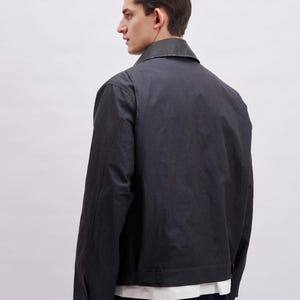 K&ouml;nnte beinhalten: Dunkelgraue Jacke mit Kragen und gekn&ouml;pften Manschetten. Die Jacke hat ein schlichtes Design und ein leicht gl&auml;nzendes Finish. Das Model tr&auml;gt ein wei&szlig;es Hemd darunter und dunkelblaue Jeans.