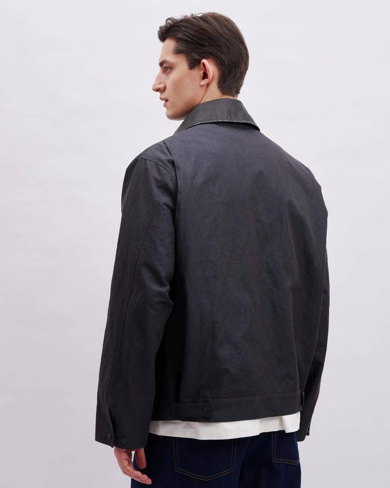 K&ouml;nnte beinhalten: Dunkelgraue Jacke mit Kragen und gekn&ouml;pften Manschetten. Die Jacke hat ein schlichtes Design und ein leicht gl&auml;nzendes Finish. Das Model tr&auml;gt ein wei&szlig;es Hemd darunter und dunkelblaue Jeans.
