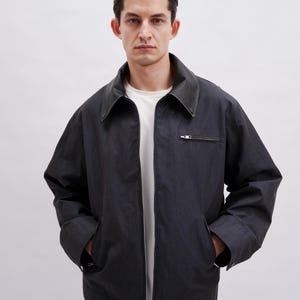 May include: Dark navy jacket with a black leather collar and a silver zipper. The jacket has two front pockets and a white t-shirt underneath. The jacket is a casual style.