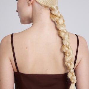 Op de afbeelding: Een vrouw met lang blond haar gevlochten in een dikke vlecht, draagt een bruin topje zonder mouwen.