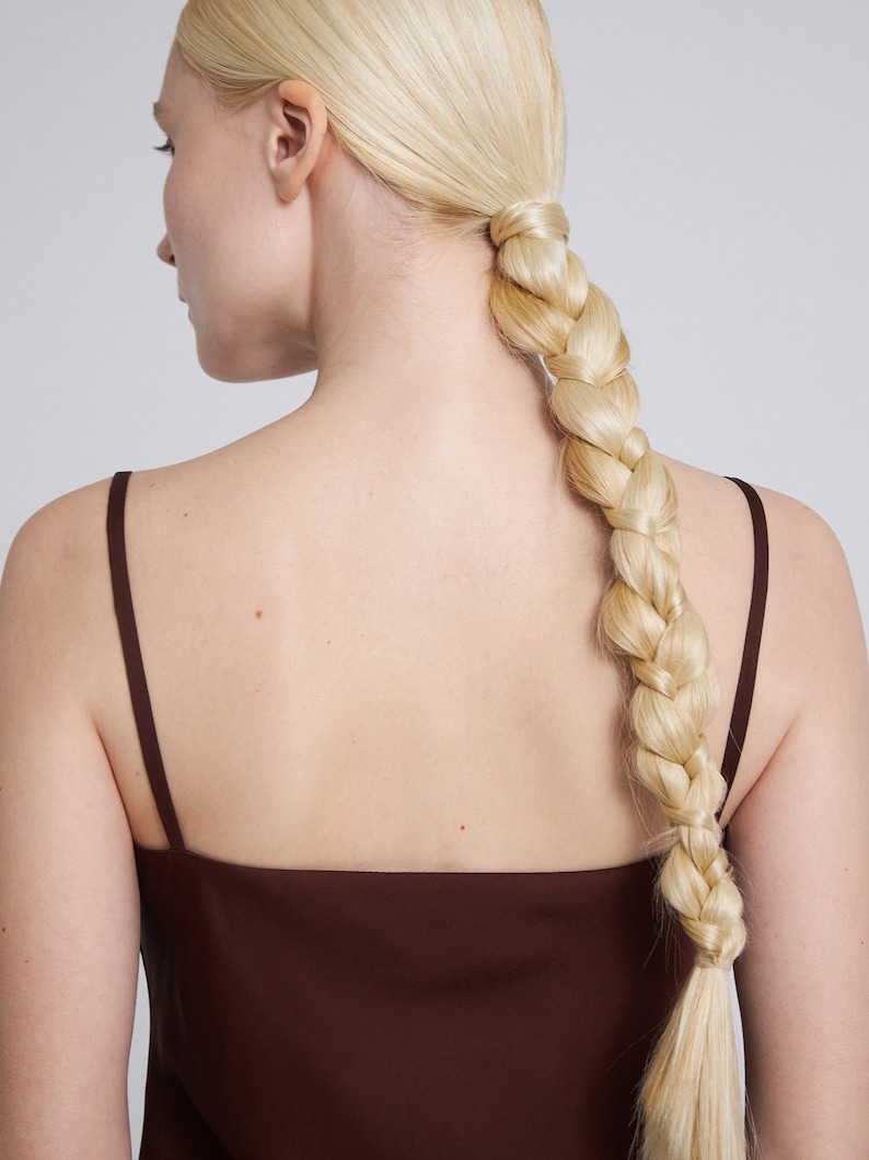 Op de afbeelding: Een vrouw met lang blond haar gevlochten in een dikke vlecht, draagt een bruin topje zonder mouwen.