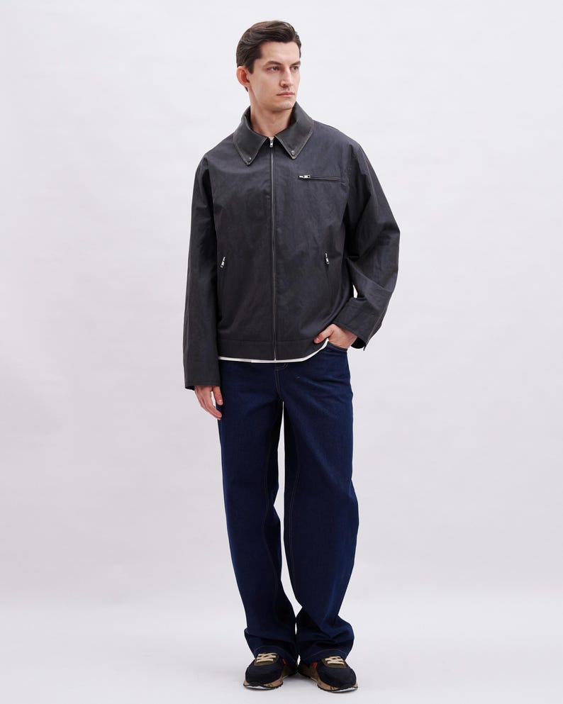 K&ouml;nnte beinhalten: Dunkelgraue Jacke mit dunkelgr&uuml;nem Kragen und Rei&szlig;verschluss. Dazu werden dunkelblaue Jeans und schwarze Sneaker getragen. Das Outfit ist l&auml;ssig und modern.