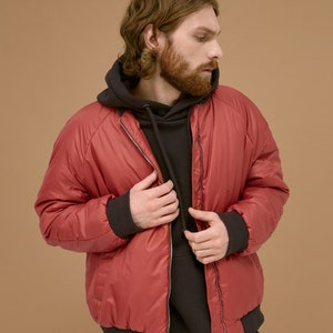May include: A red bomber jacket with a smooth finish, featuring a front zipper closure and dark charcoal ribbed cuffs and hem. The jacket is worn over a dark charcoal hooded sweatshirt with a drawstring, creating a layered look.