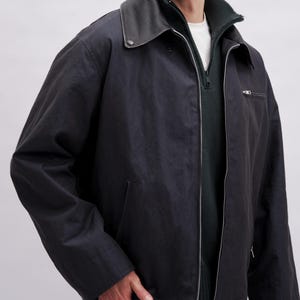 K&ouml;nnte beinhalten: Dunkelmarineblaue Jacke mit schwarzem Lederkragen und silbernem Rei&szlig;verschluss. Die Jacke hat einen Rei&szlig;verschluss vorne, eine kleine Brusttasche und zwei Seitentaschen. Das Model tr&auml;gt ein wei&szlig;es Hemd und einen gr&uuml;nen Pullover.