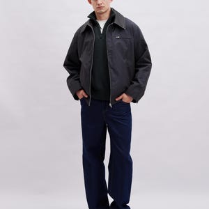 May include: A person wearing a dark gray jacket with a zipper, a dark green sweater, a white shirt, and dark blue jeans. The jacket has a collar and a small pocket on the chest. The person is wearing black and brown sneakers.