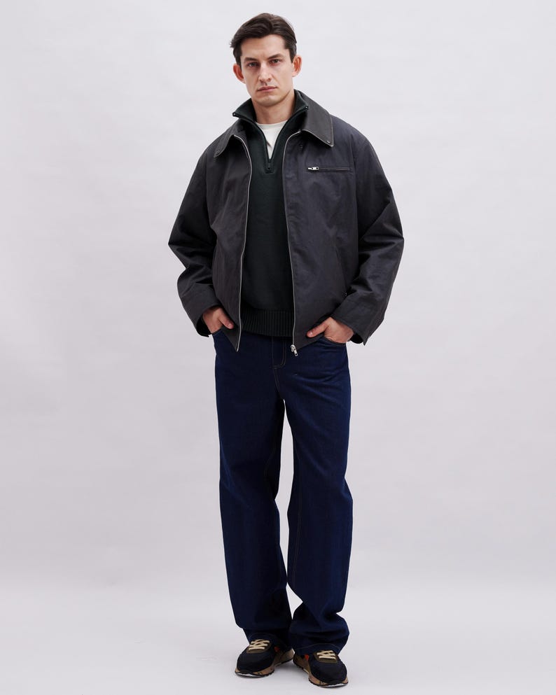 May include: A person wearing a dark gray jacket with a zipper, a dark green sweater, a white shirt, and dark blue jeans. The jacket has a collar and a small pocket on the chest. The person is wearing black and brown sneakers.