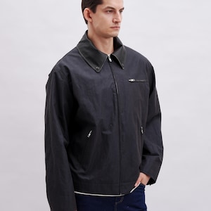 K&ouml;nnte beinhalten: Dunkelgraue Jacke mit schwarzem Kragen und Rei&szlig;verschluss. Die Jacke hat zwei Vordertaschen und eine lockere Passform. Das Model tr&auml;gt ein wei&szlig;es Hemd und dunkelblaue Jeans.