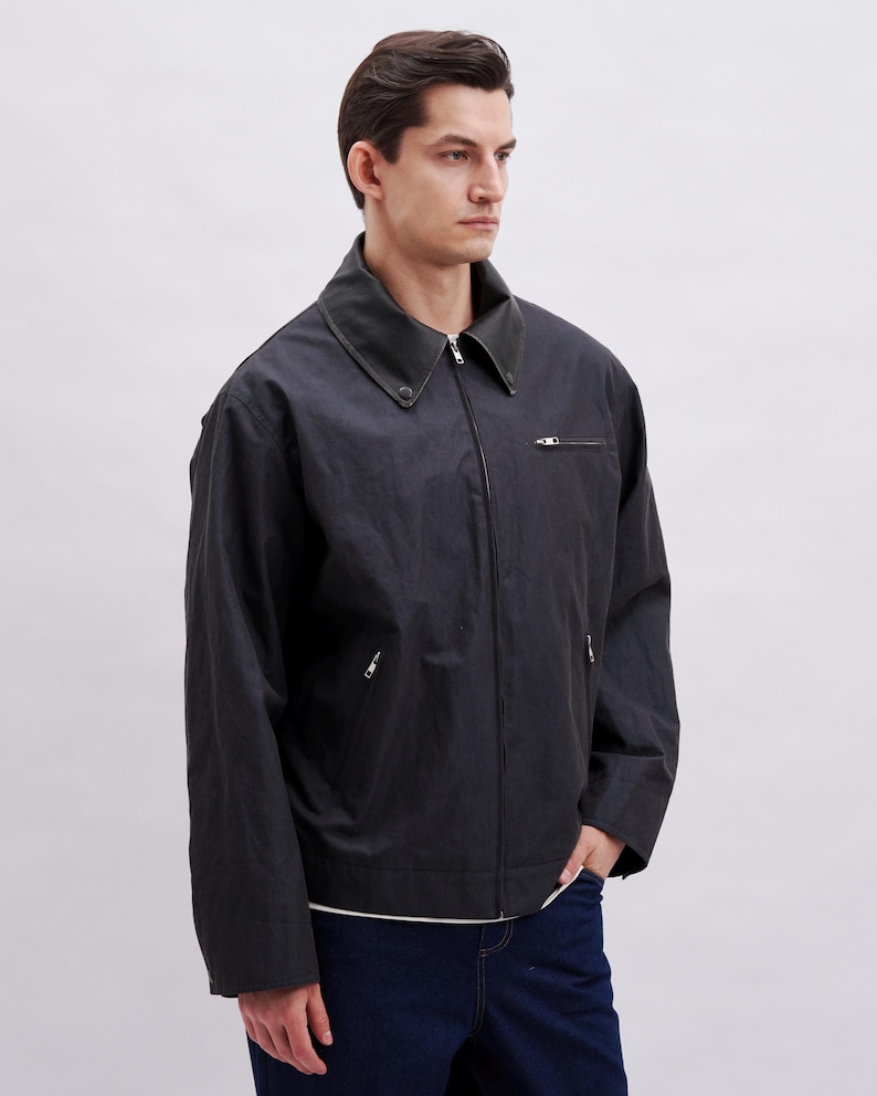 K&ouml;nnte beinhalten: Dunkelgraue Jacke mit schwarzem Kragen und Rei&szlig;verschluss. Die Jacke hat zwei Vordertaschen und eine lockere Passform. Das Model tr&auml;gt ein wei&szlig;es Hemd und dunkelblaue Jeans.