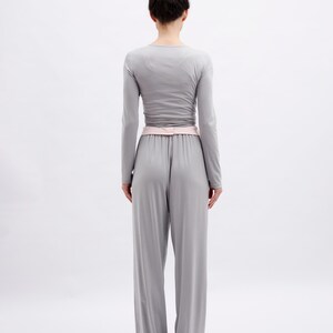 May include: A light gray long-sleeved top and matching wide-leg trousers set. The top features a fitted silhouette with long sleeves, while the trousers have a high-waisted design with a soft pink waistband detail, creating a cohesive monochromatic look.