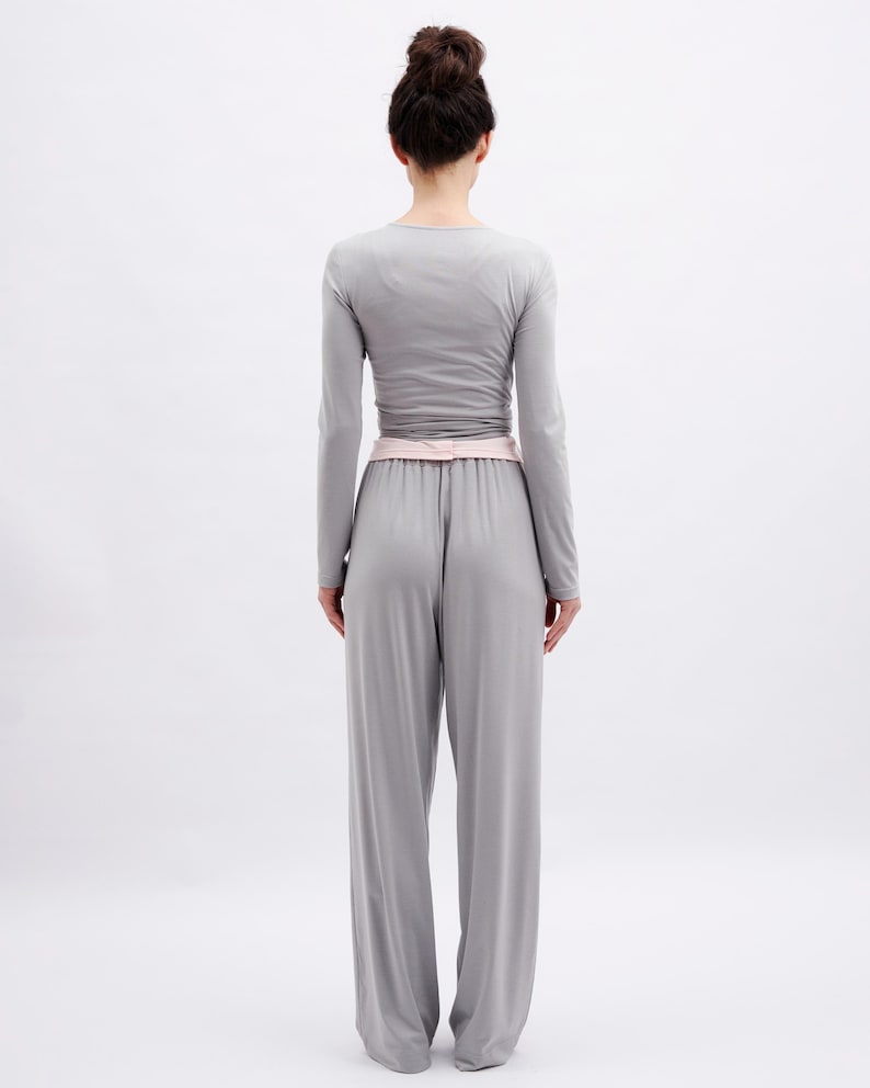 May include: A light gray long-sleeved top and matching wide-leg trousers set. The top features a fitted silhouette with long sleeves, while the trousers have a high-waisted design with a soft pink waistband detail, creating a cohesive monochromatic look.