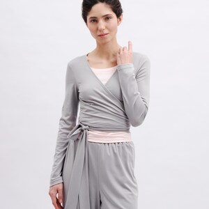 May include: A long-sleeved gray wrap top featuring a V-neckline and a side tie closure, layered over a light pink undershirt, paired with matching gray high-waisted trousers with an elastic waistband, creating a coordinated two-piece set.