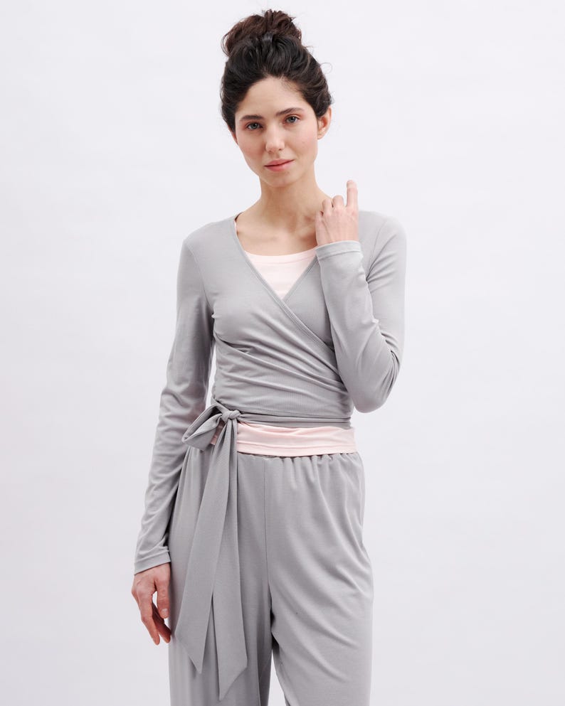 May include: A long-sleeved gray wrap top featuring a V-neckline and a side tie closure, layered over a light pink undershirt, paired with matching gray high-waisted trousers with an elastic waistband, creating a coordinated two-piece set.