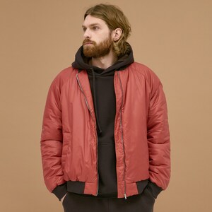 May include: A brick red bomber jacket with a smooth finish, featuring a full-length silver zipper, ribbed dark brown cuffs, and a ribbed hem. The jacket is worn over a dark brown hooded sweatshirt with a drawstring and matching dark brown pants.