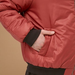 May include: A red bomber jacket featuring a smooth, slightly reflective finish, long sleeves with dark brown ribbed cuffs, and a matching dark brown ribbed hem. The jacket includes a side pocket and a vertical metal zipper closure along the front.