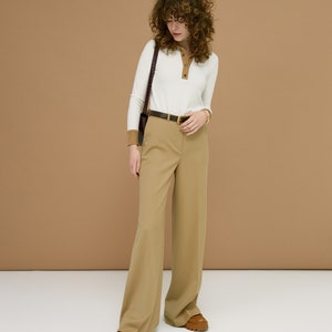 May include: A woman wearing a white long-sleeve shirt with brown trim and tan pants. The outfit includes a black belt, a brown shoulder bag, and brown shoes. The background is a solid tan color.