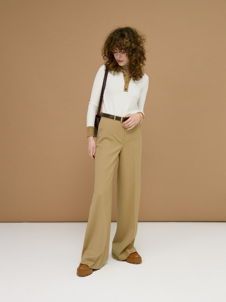 May include: A woman wearing a white long-sleeve shirt with brown trim and tan pants. The outfit includes a black belt, a brown shoulder bag, and brown shoes. The background is a solid tan color.