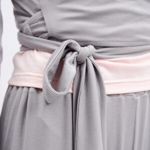 May include: A gray wrap-style top featuring long sleeves and a fabric tie at the waist, layered over a light pink undershirt. The garment is constructed from a smooth, solid-colored fabric with visible stitching along the wrap closure and hem.