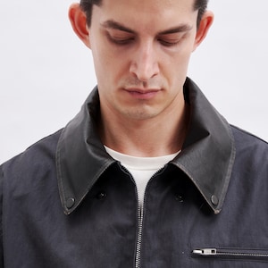 May include: A dark navy jacket with a black leather collar and a silver zipper. The jacket has a small pocket with a zipper on the chest. The person in the image is wearing a white t-shirt underneath.