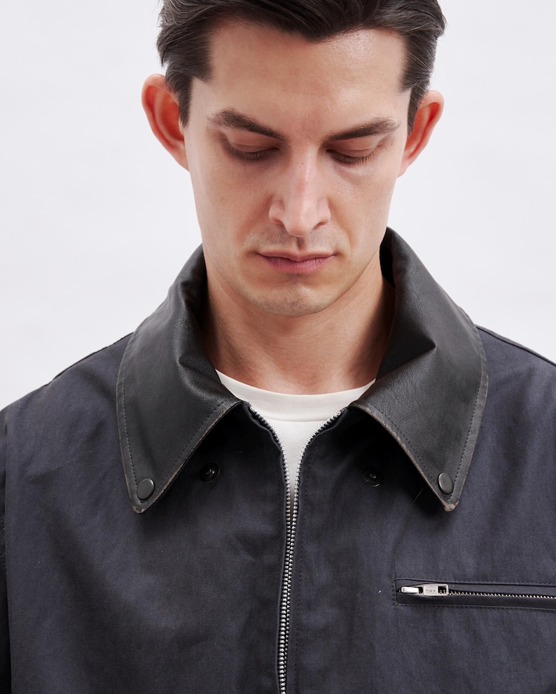 May include: A dark navy jacket with a black leather collar and a silver zipper. The jacket has a small pocket with a zipper on the chest. The person in the image is wearing a white t-shirt underneath.
