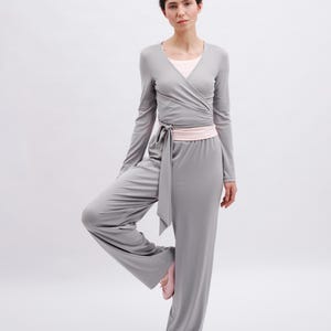 May include: A light gray long-sleeve wrap top with a tie waist and matching wide-leg trousers. The outfit is layered over a pale pink undershirt and paired with pale pink ballet slippers, creating a coordinated set in a soft, solid fabric.