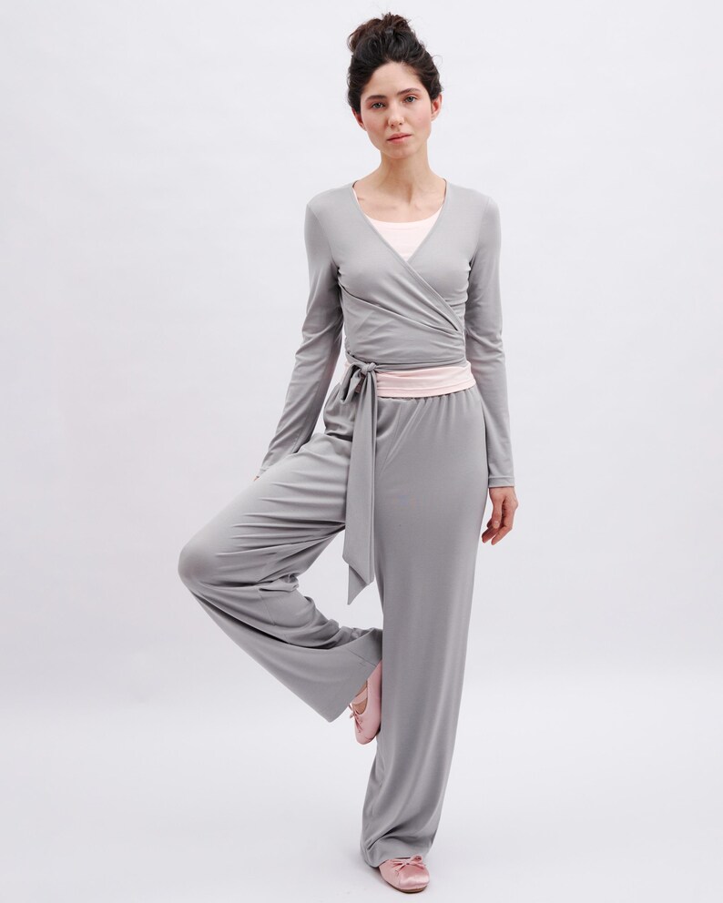 May include: A light gray long-sleeve wrap top with a tie waist and matching wide-leg trousers. The outfit is layered over a pale pink undershirt and paired with pale pink ballet slippers, creating a coordinated set in a soft, solid fabric.