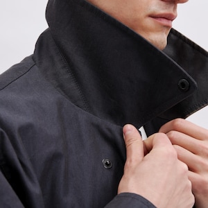 May include: A close-up of a dark gray jacket with a large, black collar. The jacket has a button closure and a zipper. The person's hands are adjusting the collar. The jacket appears to be made of a durable, possibly water-resistant material.