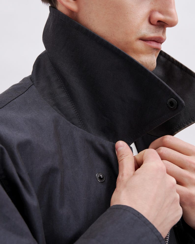 May include: A close-up of a dark gray jacket with a large, black collar. The jacket has a button closure and a zipper. The person's hands are adjusting the collar. The jacket appears to be made of a durable, possibly water-resistant material.