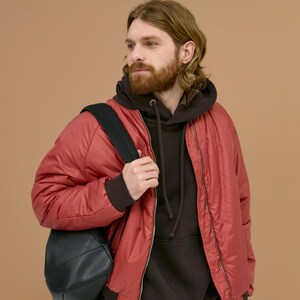 May include: A red bomber jacket with a front zipper closure worn over a dark brown hooded sweatshirt. A black backpack is carried over one shoulder. The jacket features ribbed cuffs and a smooth, slightly reflective synthetic fabric finish.