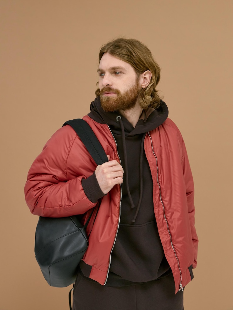May include: A red bomber jacket with a front zipper closure worn over a dark brown hooded sweatshirt. A black backpack is carried over one shoulder. The jacket features ribbed cuffs and a smooth, slightly reflective synthetic fabric finish.
