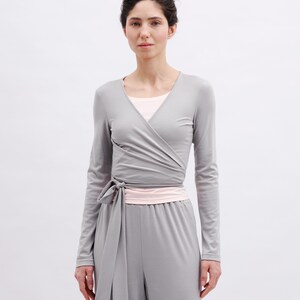 May include: A long-sleeve gray wrap top featuring a V-neckline and a side tie closure, layered over a light pink undershirt, paired with matching gray wide-leg trousers. The ensemble is constructed from a smooth, solid-colored fabric.