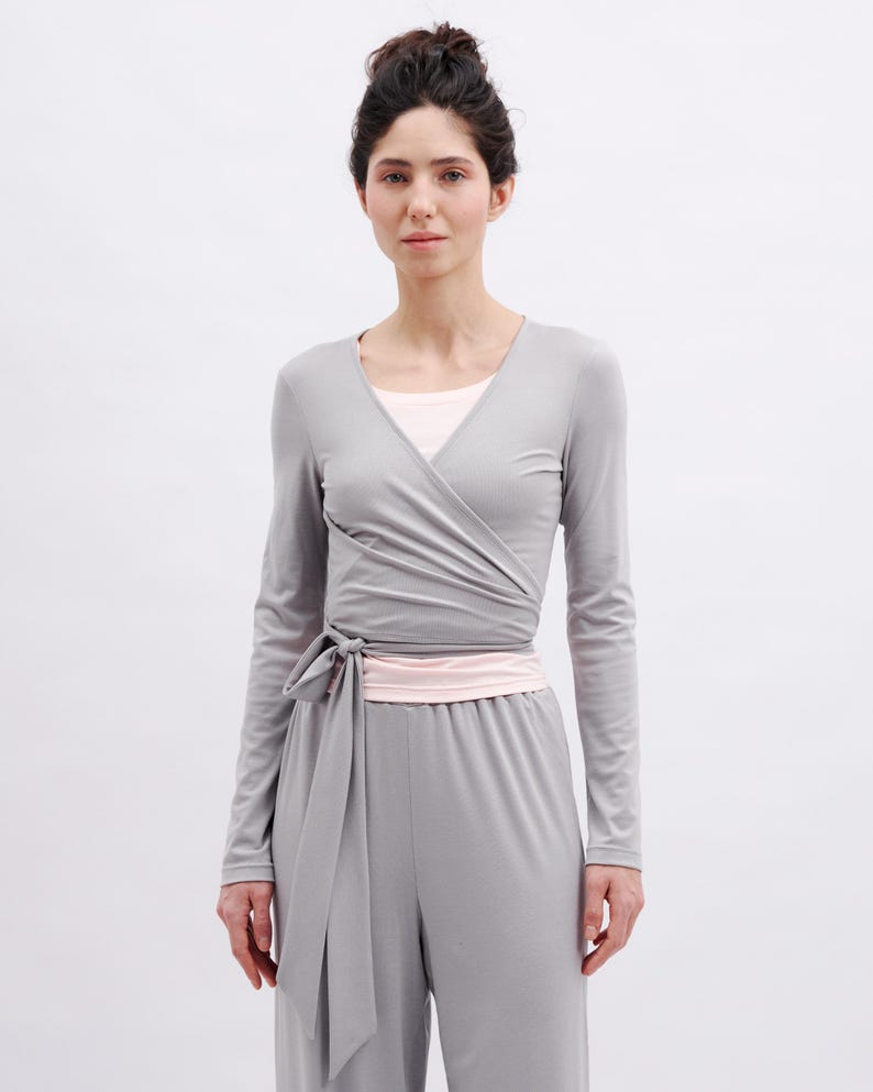 May include: A long-sleeve gray wrap top featuring a V-neckline and a side tie closure, layered over a light pink undershirt, paired with matching gray wide-leg trousers. The ensemble is constructed from a smooth, solid-colored fabric.