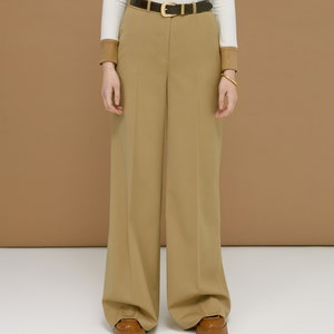 May include: Tan wide-leg trousers with a brown leather belt featuring a gold buckle. The outfit includes a white ribbed long-sleeve shirt and brown leather shoes. The background is a solid tan color.