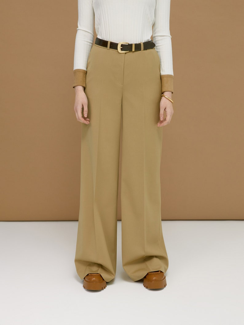 May include: Tan wide-leg trousers with a brown leather belt featuring a gold buckle. The outfit includes a white ribbed long-sleeve shirt and brown leather shoes. The background is a solid tan color.