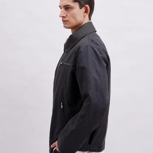 May include: A dark gray jacket with a collar and zipper closure. The jacket has a subtle sheen and a tailored fit. The model is wearing a white shirt underneath. The jacket appears to be made of a durable material.