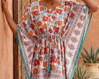 Op de afbeelding: Een witte kaftan-jurk met een levendig bloemenpatroon in oranje en rode tinten, geaccentueerd met een blauwgroene bies langs de randen. Het kledingstuk heeft een taille met trekkoord en blauwgroene kwastjes en een V-halslijn.