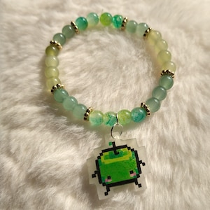 May include: A bracelet with green and gold beads, and a pixelated apple charm. The bracelet has light green, round beads with gold-coloured spacers. A pixelated green apple charm is attached. The bracelet is shown on a white, fluffy surface.