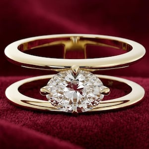 Può includere: Un anello in oro con design a doppia fascia e una pietra preziosa trasparente dal taglio ovale al centro. Le due fasce sono collegate da una piccola barra verticale, creando uno spazio aperto in cui la pietra sfaccettata è fissata a griffe.