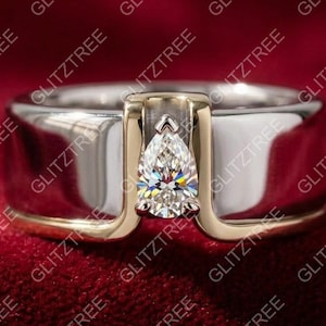 May include: A two-tone ring with a pear-shaped diamond center stone. The ring features a silver band with gold accents around the diamond setting. The ring is set against a red background.