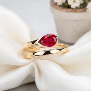May include: A gold ring featuring a single pear-shaped red gemstone set at the top. The band is smooth and polished, with the gemstone positioned horizontally. The ring is displayed on a soft, white fabric surface with a subtle sheen.