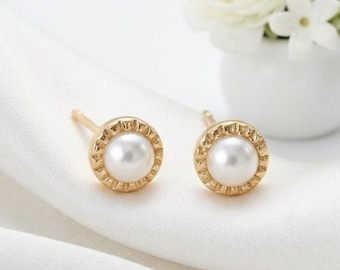May include: A pair of gold stud earrings featuring a central round white pearl surrounded by a textured, scalloped gold border. The earrings are positioned on a soft, white fabric surface, showcasing a classic design with a polished finish.