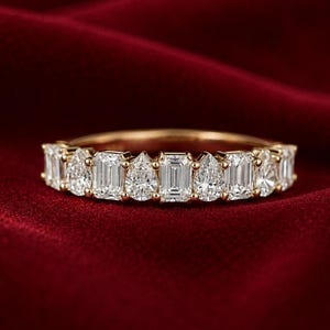 Può includere: Un anello con fascia in oro con una serie di diamanti di varie forme, tra cui smeraldo e a goccia. L'anello è posto su uno sfondo rosso intenso, che mette in risalto la brillantezza delle pietre preziose.