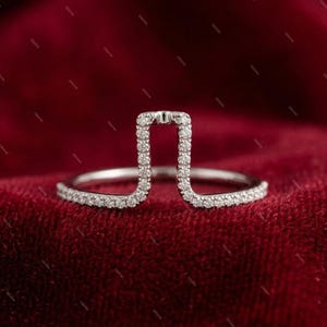 Peut inclure: Une bague en argent au design ouvert unique. La bague présente une pièce maîtresse rectangulaire ornée de petits diamants étincelants. La bande est également ornée de diamants, créant un éclat continu. La bague est posée sur un fond rouge foncé.