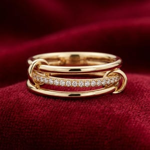 May include: A gold ring with three bands. The middle band is encrusted with small, clear gemstones. The ring has a unique design with connecting loops. The ring is set against a red background.