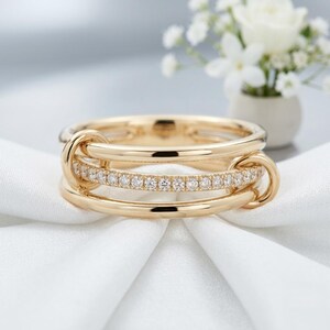 Può includere: Un anello in oro composto da tre fasce interconnesse. La fascia centrale è incastonata con una fila di piccoli diamanti rotondi e trasparenti, mentre le due fasce esterne sono in oro liscio e lucido, collegate da piccoli anelli d'oro.