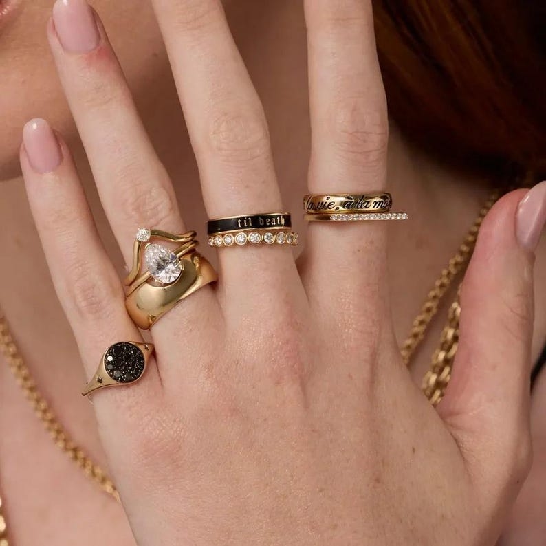 Puede incluir: Una colecci&oacute;n de anillos de oro en cuatro dedos. Un anillo tiene una piedra clara en forma de pera, otro tiene una banda negra con el texto 'til death', un tercero muestra el texto 'la vie a la mort' y un cuarto tiene una cara circular negra.