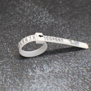 May include: White plastic ring sizer with black markings for measuring ring sizes from 4 to 17. The sizer has the text "PAT. PEND. 9101693" printed on it.