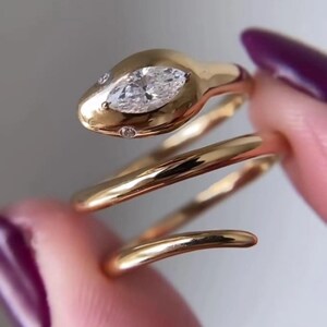 Può includere: Un anello in oro a forma di serpente con una pietra preziosa trasparente taglio marquise incastonata nella testa e due piccole pietre rotonde trasparenti come occhi. La fascia ha un design a spirale liscia e lucida.