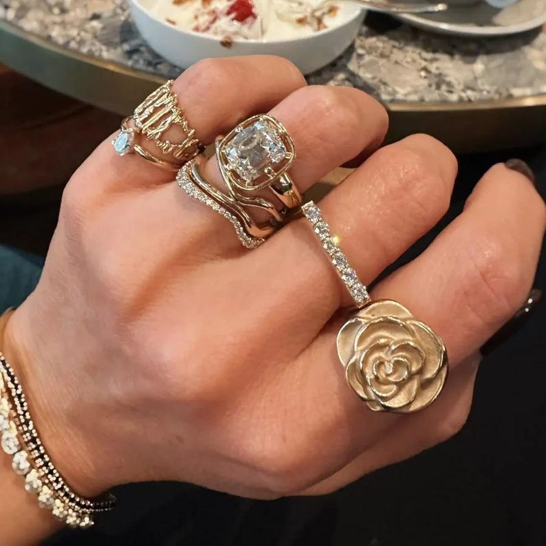 Puede incluir: Una colecci&oacute;n de anillos de oro en los dedos, que incluye un anillo central grande con una piedra clara cuadrada, una banda delicada con piedras claras peque&ntilde;as, una banda dorada texturizada y un anillo dorado con forma de flor de rosa.