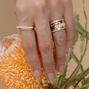 Puede incluir: Una colecci&oacute;n de anillos de oro con diversos dise&ntilde;os, incluyendo bandas con peque&ntilde;os diamantes en pav&eacute;, un anillo con tres diamantes redondos prominentes y un anillo de barra plana, todos usados en varios dedos de una sola mano.