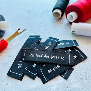 May include: A pile of black fabric labels with white text. The labels have the German phrase "ich lass' das jetzt so" on them. The labels are surrounded by spools of thread in various colors.